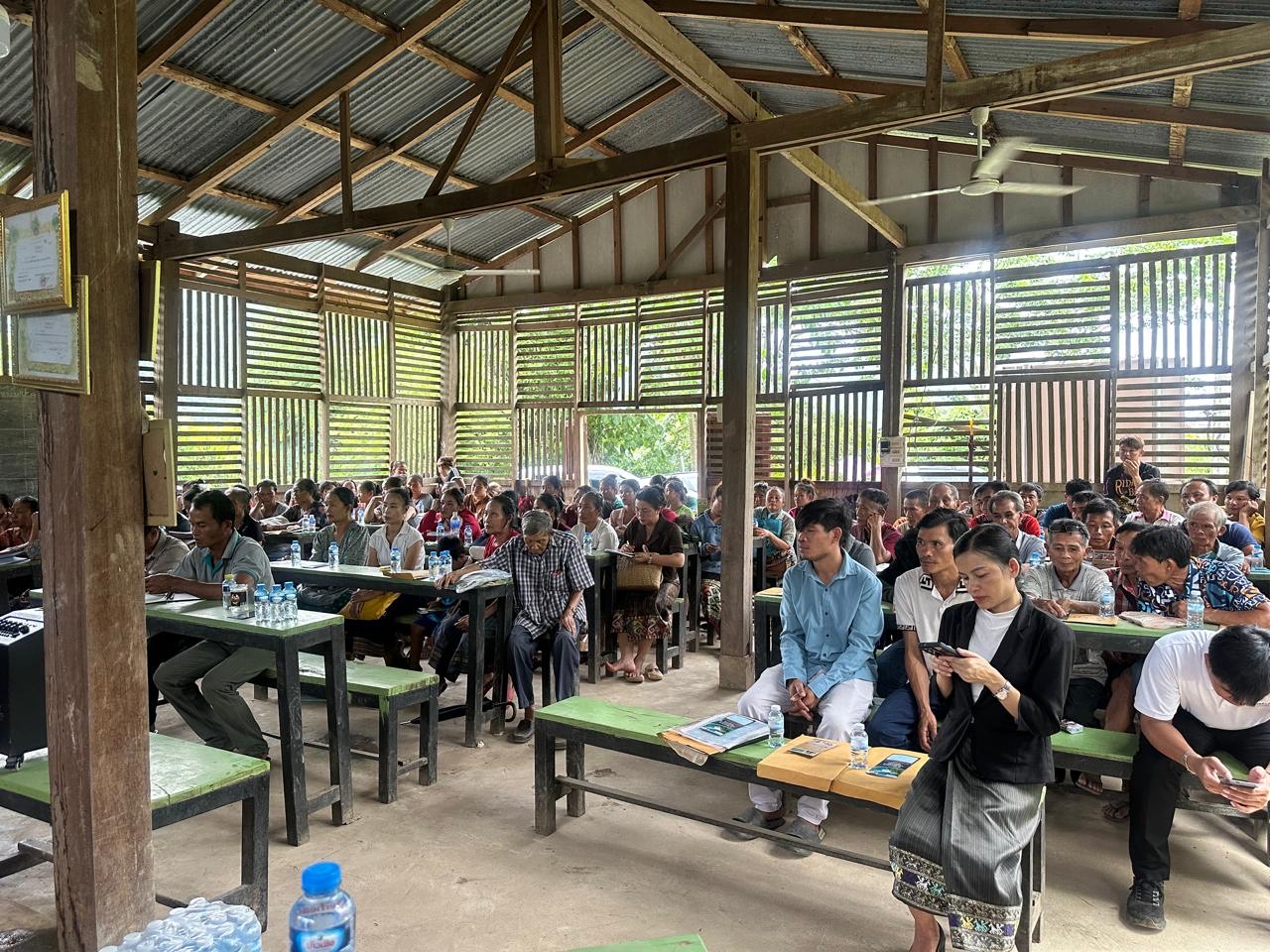 Villagers seated at benches during a resettlement information meeting in an open-sided hall